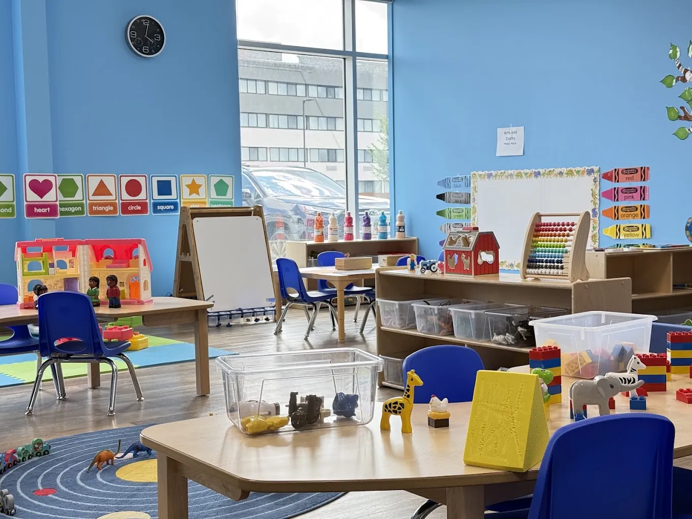 Wide view of a classroom with a dollhouse, easel, tables and shelves of toys