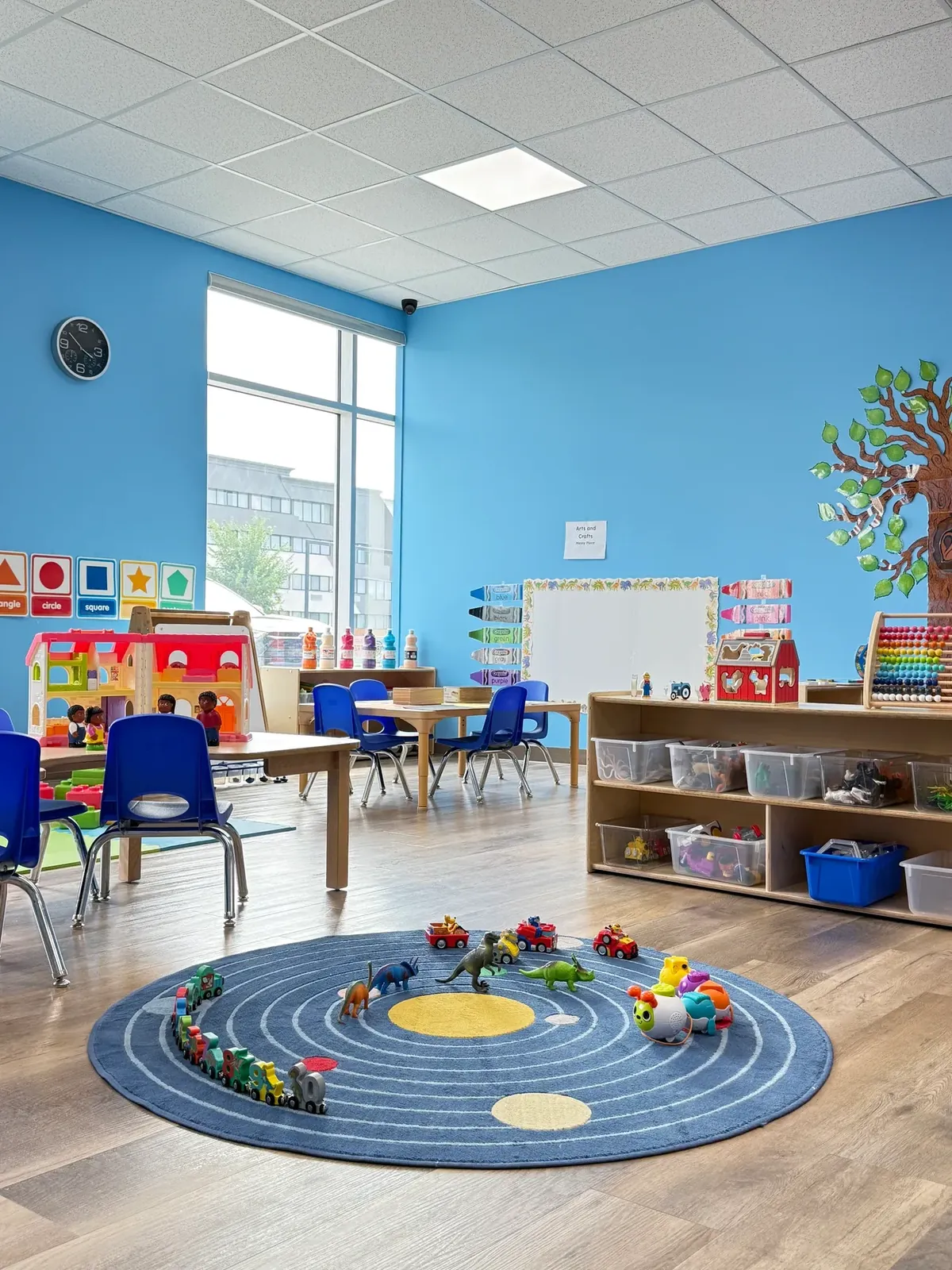Bright classroom with a round play rug, toy cars and dinosaurs, small tables and shelves of toys